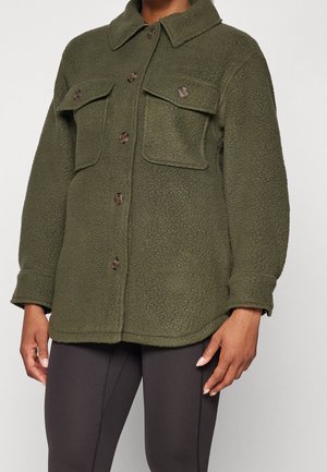 Osoba ubrana w oliwkowo-zieloną polarową kurtkę z zapinanymi na guziki kieszeniami na piersi oraz czarne legginsy, stojąca na jednolitym jasnym tle.