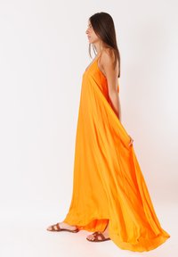 Vestito maxi arancione senza maniche realizzato in tessuto leggero, con un design fluido e scollo a V, abbinato a sandali marroni.