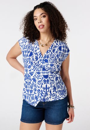 Femme aux cheveux bouclés portant un haut cache-cœur sans manches à motif bleu et blanc, un short en jean foncé et un collier avec pendentif coquillage.