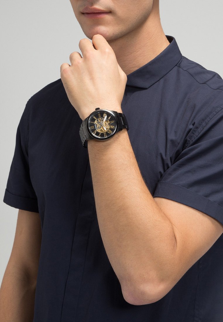 Reloj de pulsera analógico negro con acentos dorados, dial transparente y correa texturizada, llevado en la muñeca de un hombre que viste una camisa de manga corta azul marino.