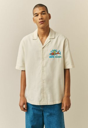 Muž v krémovej košeli s krátkym rukávom zapínanej na gombíky, s farebnou grafikou od Keitha Haringa na hrudi a v modrých džínsach.