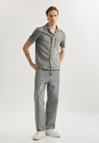 Grå denim skjorte med korte ærmer og brystlomme, parret med matchende grå denim bukser og hvide sneakers på en minimal baggrund.