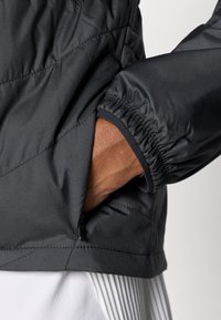 Μαύρο, καπιτονέ μπουφάν με ελαστικές μανσέτες, τσεπάκι με φερμουάρ στο πλάι και λεία υφή που αναδεικνύει το αδιάβροχο υλικό του.