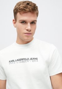 Balta kokvilnas t-kreklu ar grafisku dizainu, kurā rakstīts "KARL LAGERFELD JEANS" melnā krāsā. Ar apaļu kakla izgriezumu un īsām piedurknēm.