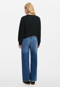 Maglione nero con maniche testurizzate, abbinato a jeans blu a gamba larga. L'outfit presenta una vestibilità comoda e uno stile informale.