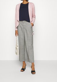 Žena v růžovém otevřeném kardiganu, tmavě modrém topu s krajkovým detailem, šedých kostkovaných širokých kalhotách, černých sandálech na podpatku a držící bílou tkanou kabelku.