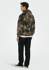 Veste à capuche en polaire au motif camouflage sombre, avec une coupe décontractée, des poignets élastiques et une capuche à cordon. Portée avec un pantalon cargo noir.