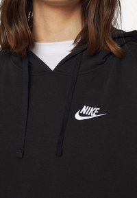 Zwarte hoodie met een wit Nike-logo op de linkerkant van de borst, voorzien van een koord in de capuchon en een zachte, gladde textuur. Gelaagd met een wit shirt eronder.