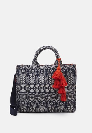 Borsa in tessuto intrecciato con un motivo geometrico blu e bianco, dotata di due manici corti e un accento di nappina rossa. Include una tracolla removibile.