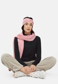 Różowa dziergana opaska na głowę i szalik, czarna bluzka z długim rękawem, beżowe spodnie z szerokimi nogawkami o fakturze oraz białe trampki, siedząca w pozycji z nogami skrzyżowanymi.
