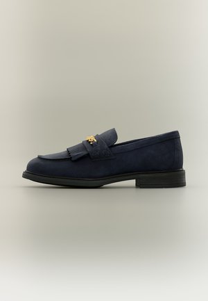 Mocassin en daim bleu foncé avec semelle noire et petit talon, avec une lanière à franges et un petit emblème décoratif doré sur le dessus.