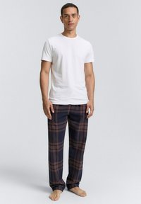 Witte crew neck t-shirt gecombineerd met marineblauwe en bruine geruite pyjamabroek, met een relaxte pasvorm en zachte stof. Model staat op blote voeten.