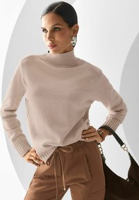 Frau mit dunklen Haaren in beigem Rollkragenpullover und braunen Hosen, die mit der Hand auf der Hüfte posiert und eine braune Handtasche hält.