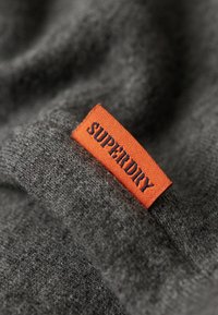 Una tela gris con una textura suave presenta una etiqueta naranja cosida, que muestra la palabra "SUPERDRY" en azul marino.