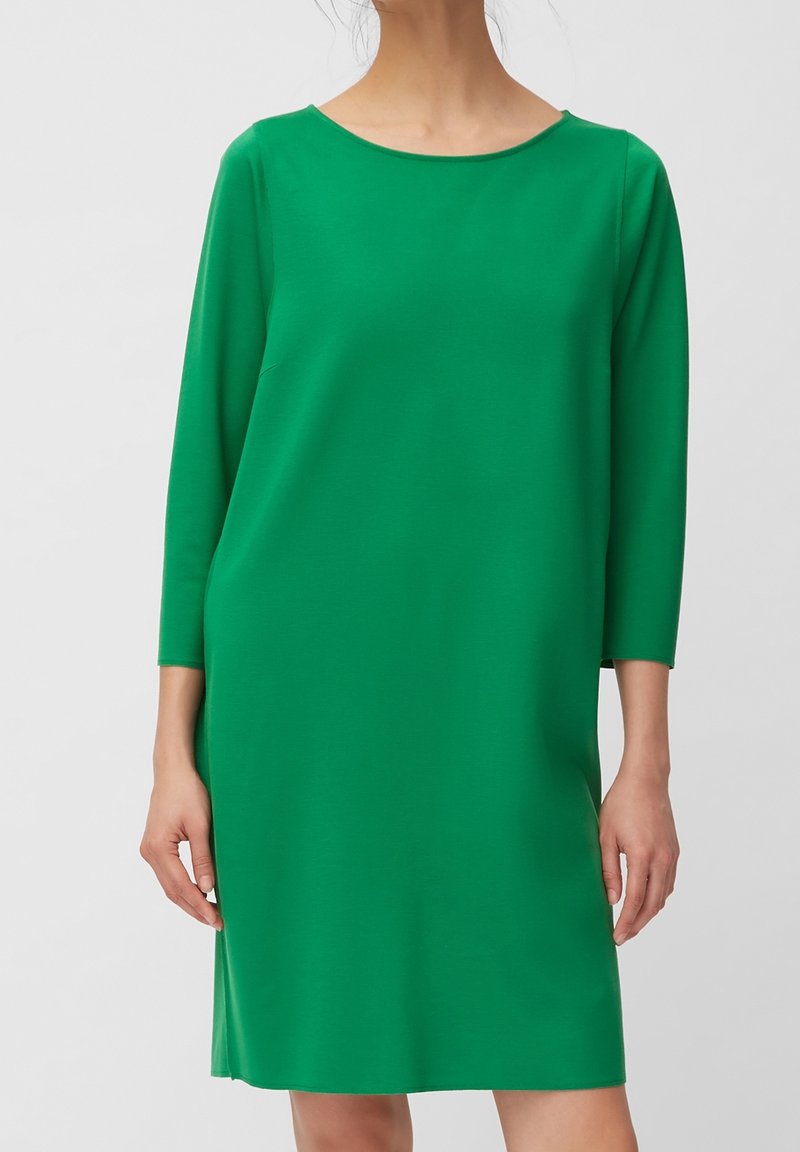 Femme portant une robe verte unie longueur genou avec des manches trois-quarts et un col rond sur un fond neutre.