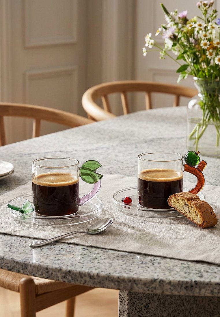 Dwie filiżanki czarnej kawy na szklanych spodkach z ozdobnymi uchwytami, na jednym spodku biscotti, łyżeczka i wazon z kwiatami na kamiennym stole.