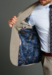 Chaqueta de traje beige claro con un forro azul con patrón de cachemira. Cuenta con un pequeño bolsillo y un cierre de botón en el interior.