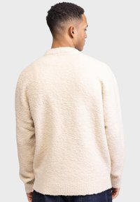 Cremefarbener, strukturierter Pullover mit lockerer Passform, der einen Schalkragen und gerippte Bündchen aufweist. Das Material wirkt weich und gemütlich.
