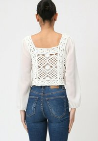 Top blanc court avec un dos en dentelle, manches longues transparentes et décolleté carré, porté avec un jean en denim bleu taille haute.