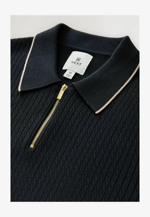 Marineblå ribbet polo skjorte med hvid kant ved kraven og en guldbelysning, med et mærke der angiver mærket og størrelse M.