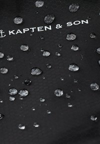 Czarny heksagonalny materiał teksturowany z kroplami wody, z białym logo "KAPTEN & SON".