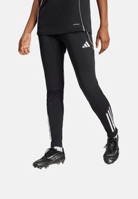 Zwarte sportleggingen en een zwart shirt met witte accenten. De legging heeft drie witte strepen aan elke kant. De schoenen zijn zwart.