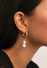 Gouden hoepeloorbel met twee witte parels die hangen, gedragen in het linkeroor van een persoon met donker haar en een gemiddelde huidskleur.