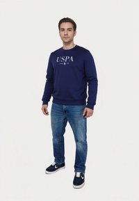 Námořnická modrá mikina s bílým textem "USPA", kulatým výstřihem a dlouhými rukávy. V kombinaci s světle modrými džíny a námořnickými teniskami s bílými akcenty.