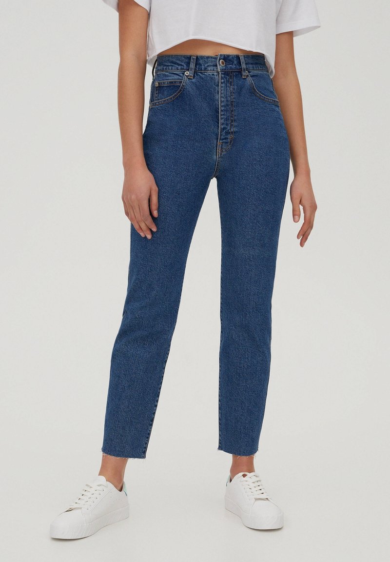 Jean en denim bleu taille haute avec une jambe droite, doté d'un ourlet effiloché. Assorti à des baskets blanches à lacets et à une chemise blanche courte.