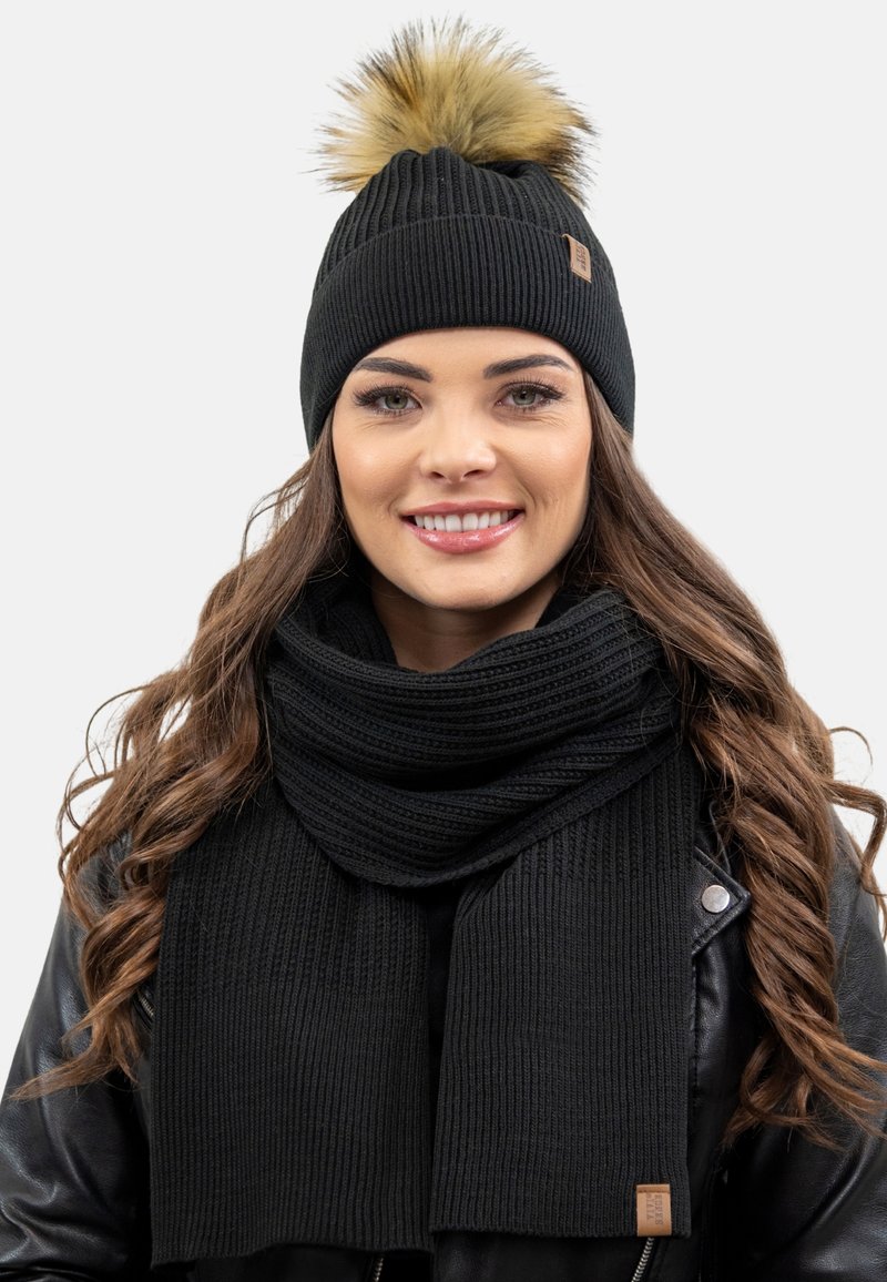 Cappello di lana nera lavorato a maglia con pompon in pelliccia sintetica e sciarpa a coste abbinata, entrambi caratterizzati da un motivo testurizzato, con un accento in giacca di pelle.