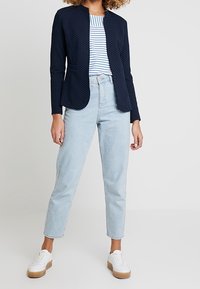 Granatowa marynarka w grochy z kołnierzem stójką, zestawiona z niebiesko-białym paskowanym topem i jasnoniebieskimi jeansami o prostych nogawkach, uzupełniona białymi trampkami.