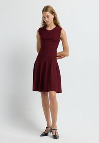 Robe sans manches bordeaux en tissu texturé, corsage ajusté, jupe évasée et plis. Portée avec des escarpins noirs à bout pointu.