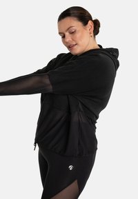 Felpa sportiva nera con maniche corte e trasparenti, schiena parzialmente trasparente e cappuccio con coulisse. Abbinata a leggings neri opachi con dettagli in rete.