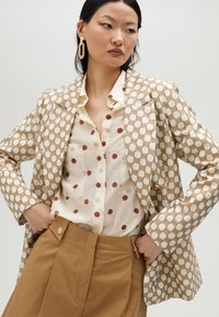 Beige mönstrad kavaj med brunt geometriskt mönster, lager på lager över en krämfärgad skjorta med röda prickar, i kombination med beige högmidjade byxor.