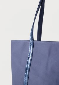 Sac fourre-tout en tissu bleu avec deux poignées, dont une décorée de sequins bleus brillants sur toute sa longueur, sur un fond blanc uni.