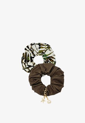 Kobieta o długich brązowych włosach, ubrana w bezramkowy top w tropikalny wzór, ciemnobrązowe doły, pasującą gumkę do włosów i gumkę na nadgarstek, uśmiechająca się.