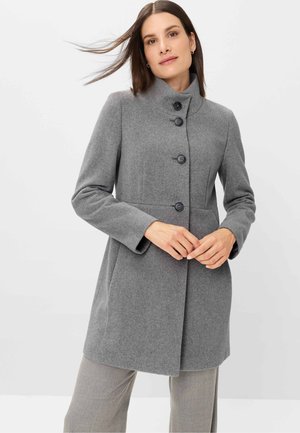 Woman with straight brown hair wearing a buttoned grey wool coat and light grey pants, standing against a plain white background.