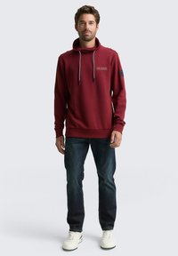 Bordeaux hoodie met trekkoord, voorlogo en zijzakken, gecombineerd met donkere jeans en witte sneakers. Zachte, gestructureerde stof.