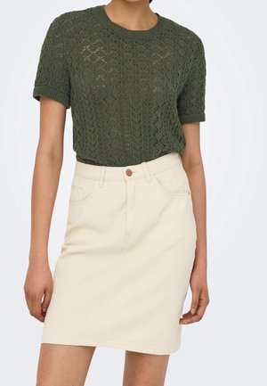 Vrouw draagt een donkergroene, gebreide top met korte mouwen, ingestopt in een beige, hooggetailleerde rok met voorzakken en knoopsluiting.