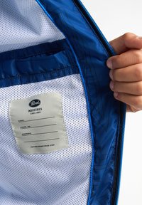 Interiorul jachetei albastre este prevăzut cu căptușeală din plasă, un etichetă din material cu numele brandului și detalii, și un margine netedă cu fermoar.