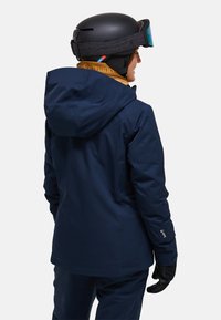 Marineblaue wasserfeste Skijacke mit Kapuze, mit elastischen Bündchen, regular Fit und glatter Textur. Getragen mit einem schwarzen Helm und Handschuhen.