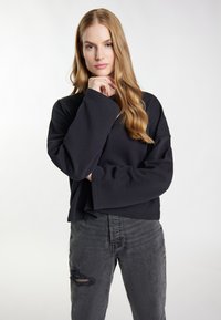 Schwarzer, gekürzter Pullover mit weiten Ärmeln, aus strukturiertem Stoff. Kombiniert mit grauen, zerrissenen Jeans für einen lässigen Look.