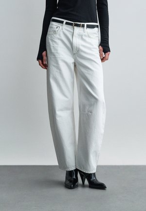 Osoba oblečená v bielych džínsach s vysokým pásom a širokými nohavicami, s čiernym opaskom, čiernymi čižmami s ihličkovými podpätkami a čiernym dlhým rukávom.