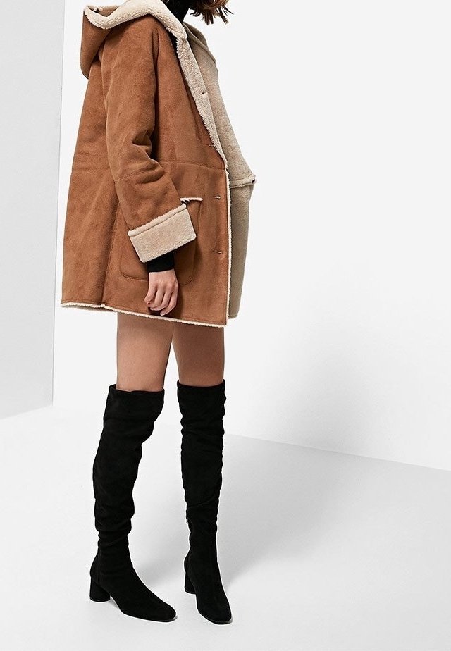 Chaqueta de shearling marrón con un forro beige, diseño oversized, mangas largas y bolsillos frontales, combinada con botas negras hasta el muslo.