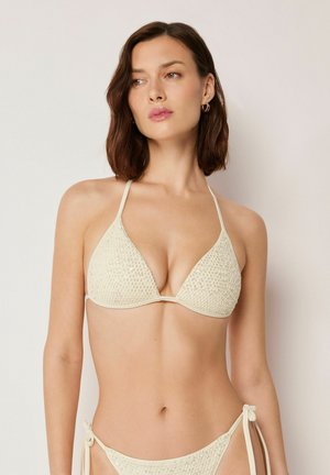 Kvinde med skulderlangt brunt hår iført en cremefarvet, tekstureret bikini med halterneck-top og sider med bindebånd, stående foran en ensfarvet baggrund.