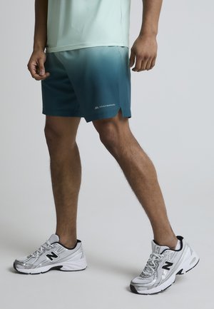 Homme portant un short dégradé bleu clair et des chaussures de course New Balance argentées, debout devant un fond uni gris clair.