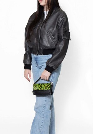 Femme portant une veste en cuir noire et un jean bleu clair, tenant un petit sac à main noir avec un rabat imprimé léopard vert.