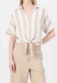 Camicia a maniche corte in lino con righe verticali beige e bianche, chiusura frontale con bottoni e dettaglio in vita annodata. Abbinata a pantaloni beige a vita alta.