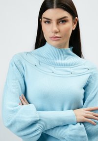 Hellblauer Pullover mit einem hohen gerippten Kragen, strukturierten Strickärmeln und dekorativen Details an den Schultern. Weiches Material und lockere Passform.