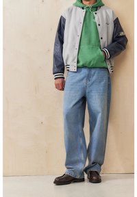 Šedá a námořnicky modrá univerzitní bunda přes zelenou mikinu s kapucí, doplněná volnými modrými džínami a hnědými botami, s kontrastními pruhy a texturami.